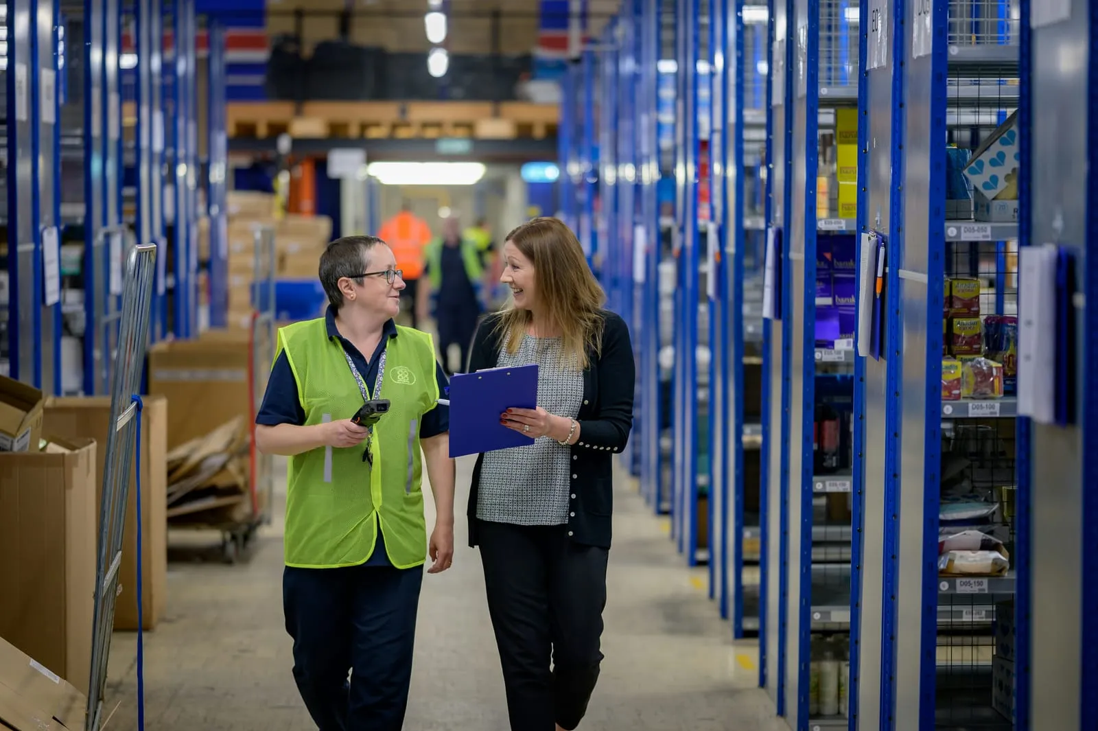 Two colleagues walking down a warehouse aisle between stocked shelving, one holding a clipboard
